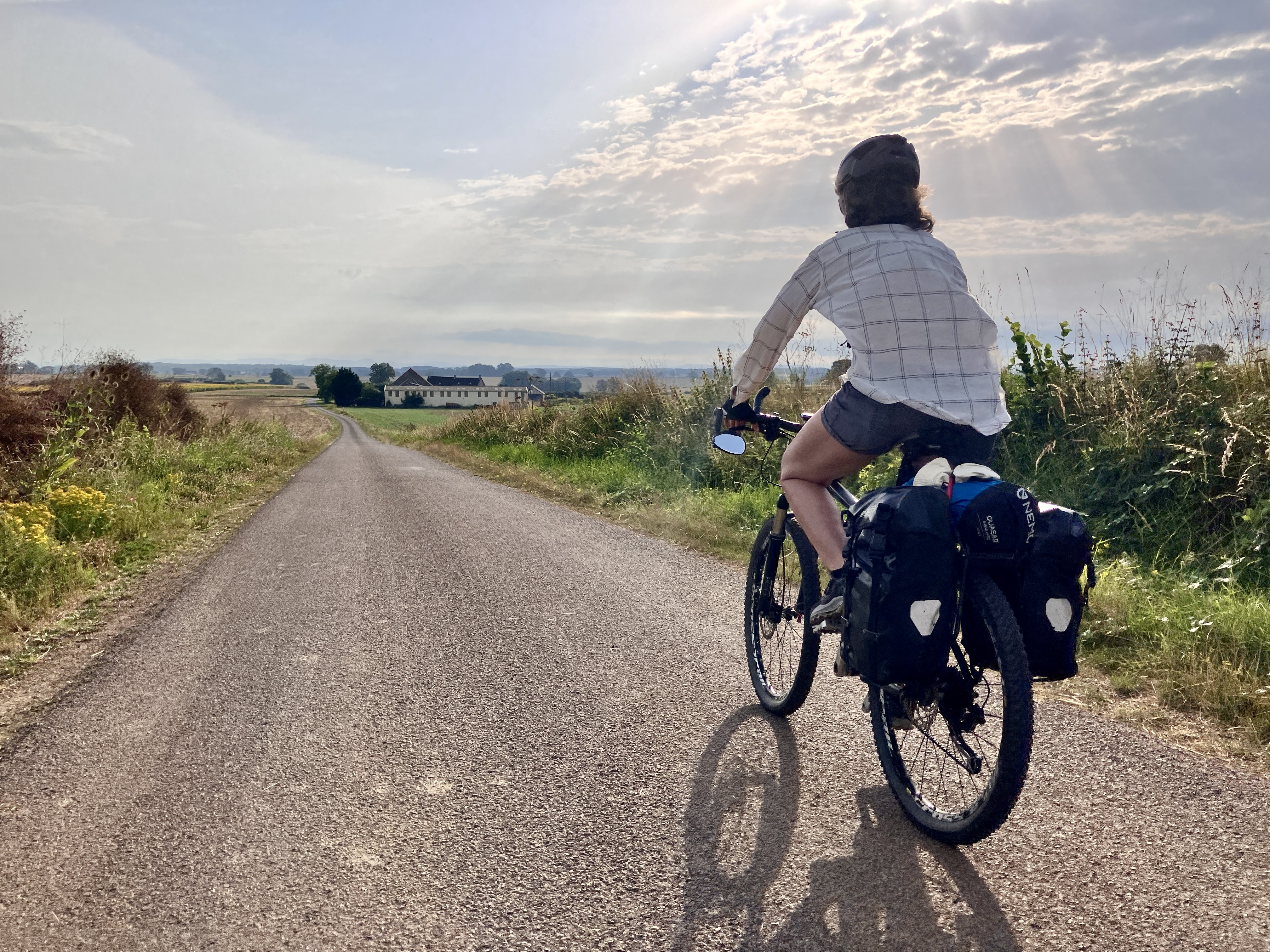 Photo d’une personne à vélo sur une route de campagne avec le soleil perçant à travers les nuages.